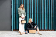 Zwei Frauen mit stilvollen, modernen Tragetaschen vor einer blau-schwarzen Wand. Eine Frau steht und hält eine kleinere Tasche, während die andere sitzt und eine größere Tasche neben sich hat. Frontale Perspektive.