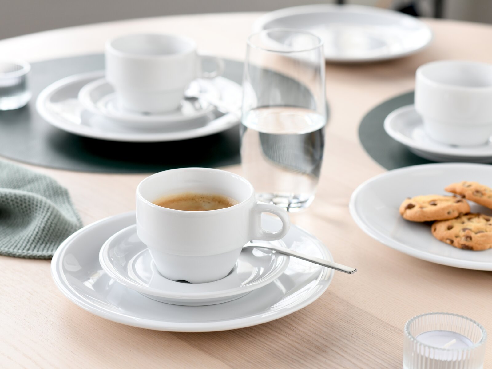 Weiße Espressotasse mit Untertasse und kleinem Löffel auf hellem Holztisch, im Hintergrund Wasserglas, weiteres Geschirr und Kekse; Perspektive: leicht erhöhte Frontansicht