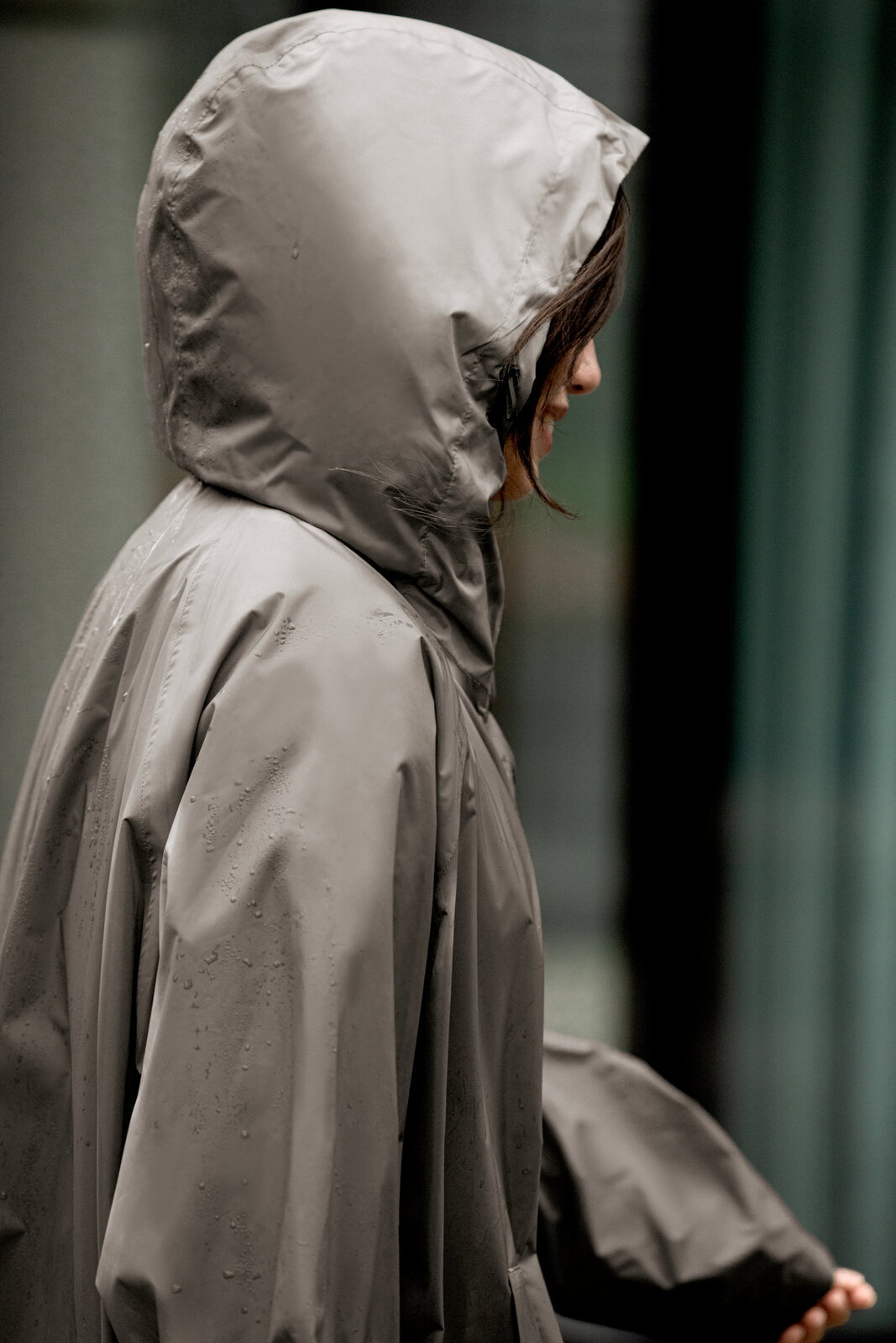 Seitenansicht einer Person in grauem, wasserdichtem Regenponcho mit Kapuze, mit sichtbaren Regentropfen auf dem Material
