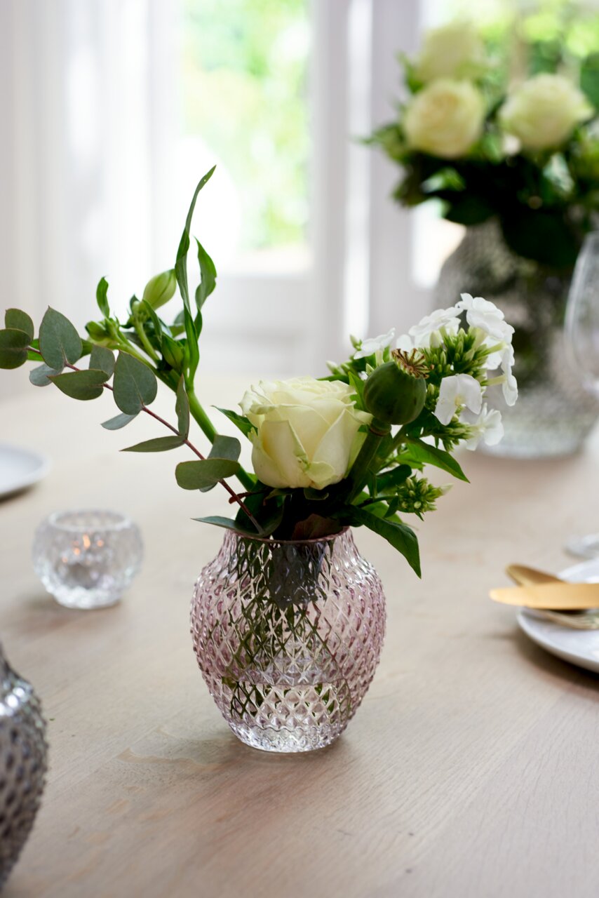 Kristallvase mit rautenförmigem Muster, gefüllt mit weißen Rosen und grünen Blättern, auf einem Tisch in einem hellen Raum, seitliche Perspektive.