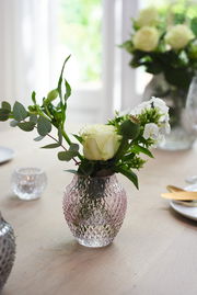 Kristallvase mit rautenförmigem Muster, gefüllt mit weißen Rosen und grünen Blättern, auf einem Tisch in einem hellen Raum, seitliche Perspektive.