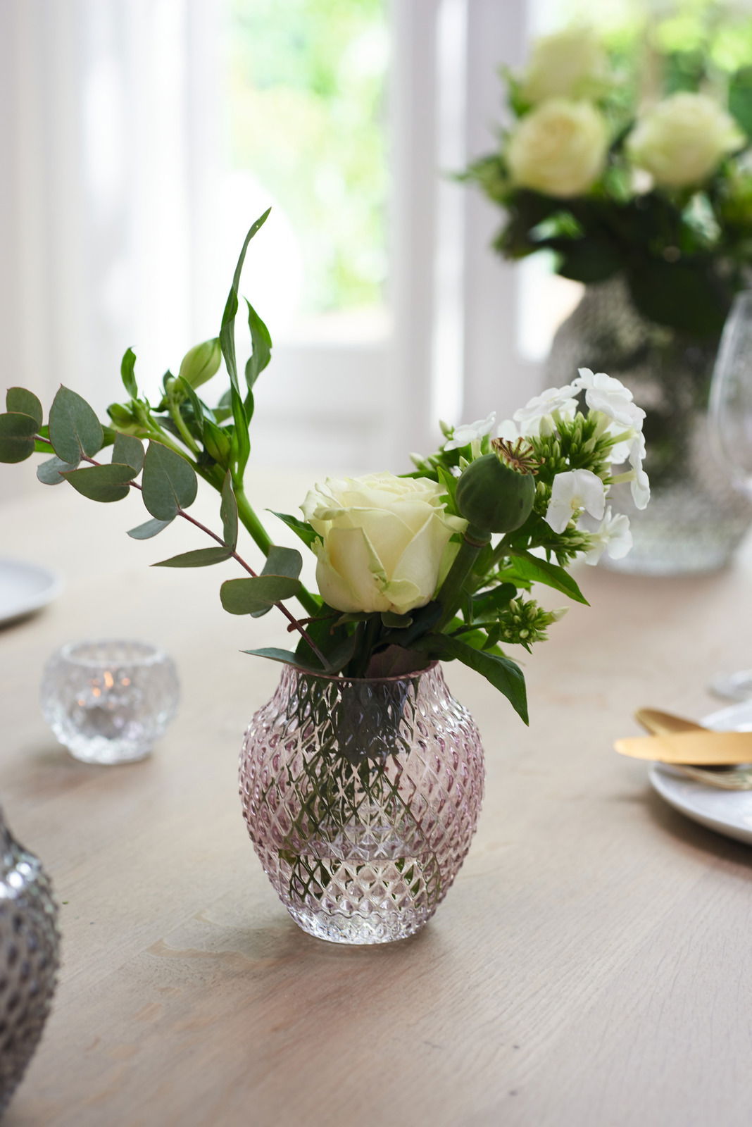 Kristallvase mit rautenförmigem Muster, gefüllt mit weißen Rosen und grünen Blättern, auf einem Tisch in einem hellen Raum, seitliche Perspektive.