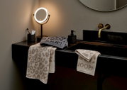 Badkamer met stijlvol opgestalde douchehanddoeken met luipaardprint en JOOP!-logo, op een wastafel naast een verlichte spiegel en een zeepdispenser, gezien vanuit een zijdelingse hoek.
