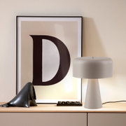Weiße Tischlampe mit zylindrischem Schirm und konischem Fuß auf einem Sideboard, daneben gerahmtes Poster mit großem Buchstaben „D“; frontale Perspektive auf das Produkt.