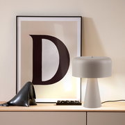 Weiße Tischlampe mit zylindrischem Schirm und konischem Fuß auf einem Sideboard, daneben gerahmtes Poster mit großem Buchstaben „D“; frontale Perspektive auf das Produkt.