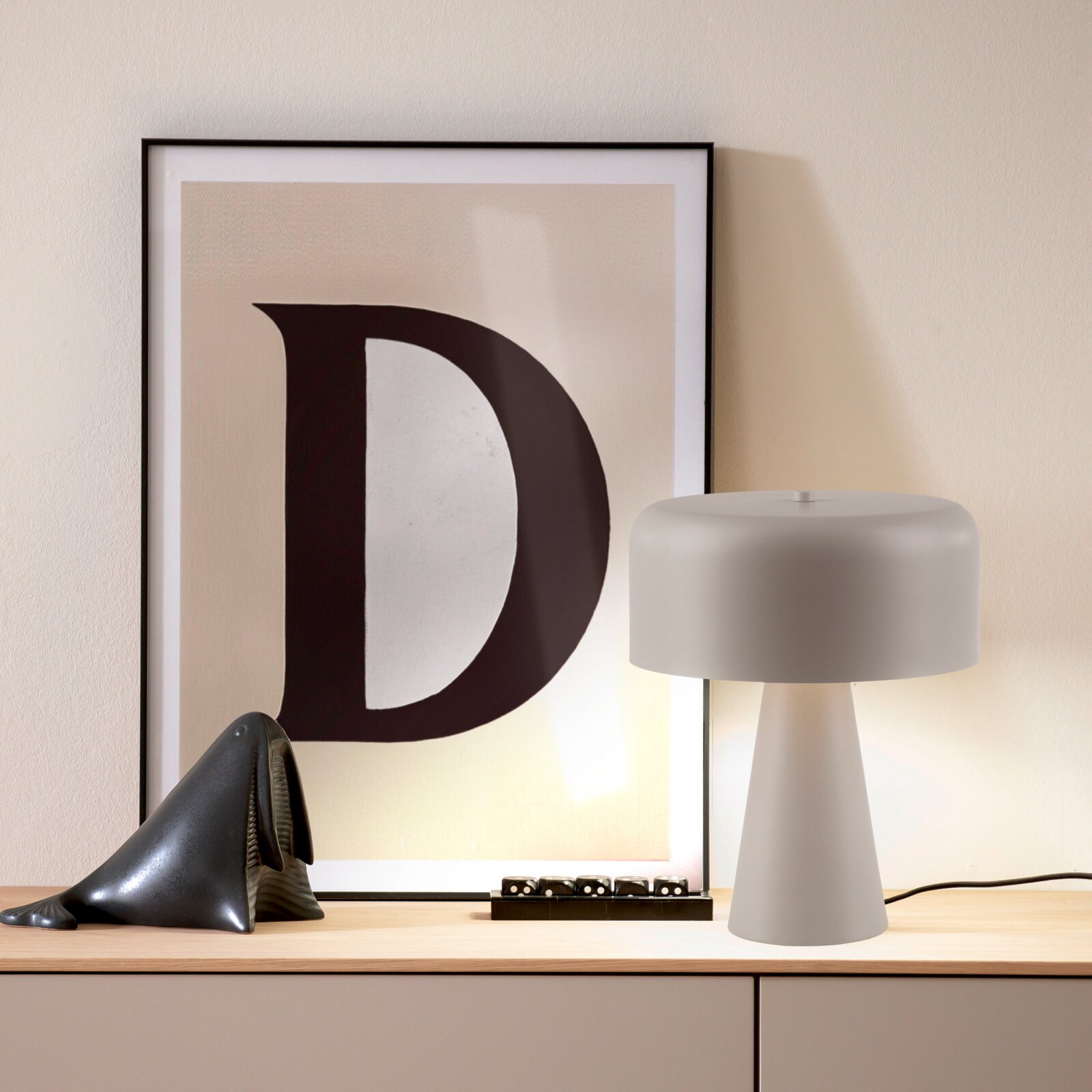 Schöner Wohnen Tischleuchte PENTO Weiße Tischlampe mit zylindrischem Schirm und konischem Fuß auf einem Sideboard, daneben gerahmtes Poster mit großem Buchstaben „D“; frontale Perspektive auf das Produkt.