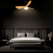 Moderner Deckenventilator mit integrierter LED-Leuchte, drei Holzflügeln und schwarzem Gehäuse über einem Bett im Schlafzimmer, Ansicht von unten in leichter Untersicht (Frontalperspektive)