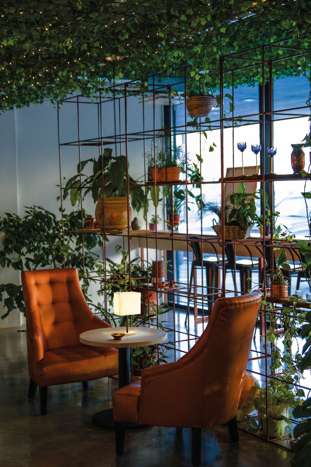 Tischleuchte Kate auf einem kleinen runden Tisch zwischen zwei orangefarbenen Sesseln, umgeben von Pflanzen in einem gemütlichen Raum.