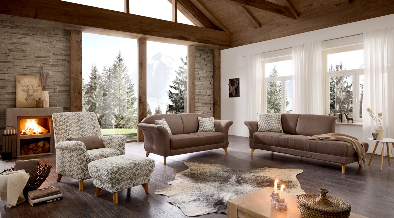 Gemütliches Wohnzimmer mit zwei braunen Sofas und einem gemusterten Sessel, Blick aus der Ecke des Raumes mit Kamin und großen Fenstern mit Bergblick.