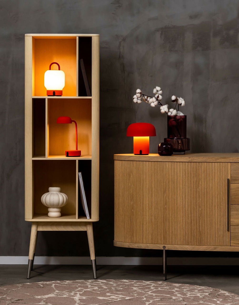 Ein stilvolles Wohnzimmer mit einem modernen Holzregal und einer Kommode, dekoriert mit drei orangefarbenen Lampen in verschiedenen Designs. Die Lampen strahlen ein warmes Licht aus und schaffen eine gemütliche Atmosphäre. Perspektive: Frontalansicht.