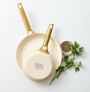 Zwei cremefarbene Bratpfannen mit goldenen Griffen, von oben fotografiert, neben frischen Kräutern und Gewürzen.