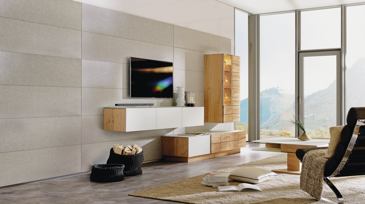 Moderne Wohnwand ohne Beleuchtung in einem hellen Wohnzimmer mit großen Fenstern, bestehend aus hängenden und stehenden Elementen in Holz- und Weißtönen, seitliche Perspektive.