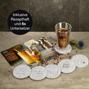 Set aus sechs runden Untersetzern mit verschiedenen Gravuren, präsentiert mit einem Glas auf einem Untersetzer und einem Rezeptheft im Hintergrund. Perspektive von oben.