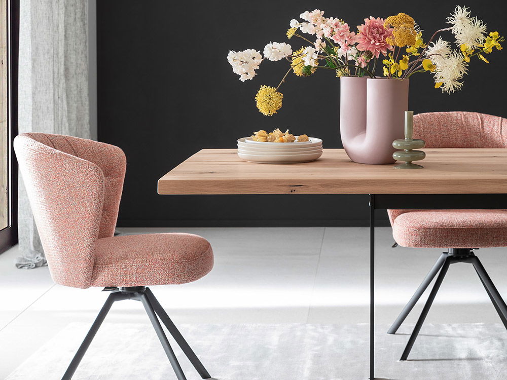 Holztisch mit rosa Stühlen, bunter Blumenvase und grauen Teppich vor dunkler Wand.