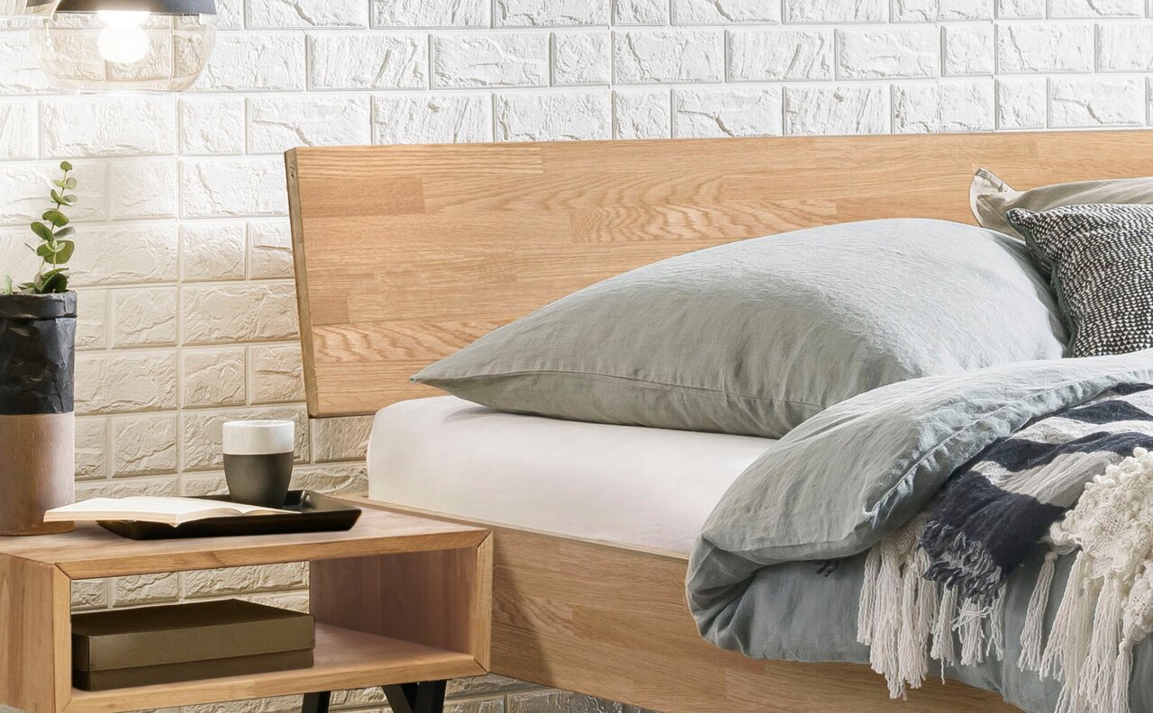 Houten bedframe met hoofdeinde van licht hout, gezien vanaf de zijkant, gedecoreerd met grijs beddengoed en kussens, met daarnaast een nachtkastje met een plant en een boek.