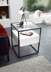 Moderner Beistelltisch mit Glasplatte und schwarzem Metallrahmen, von oben fotografiert. Auf dem Tisch stehen eine Lampe und ein Notizbuch.
