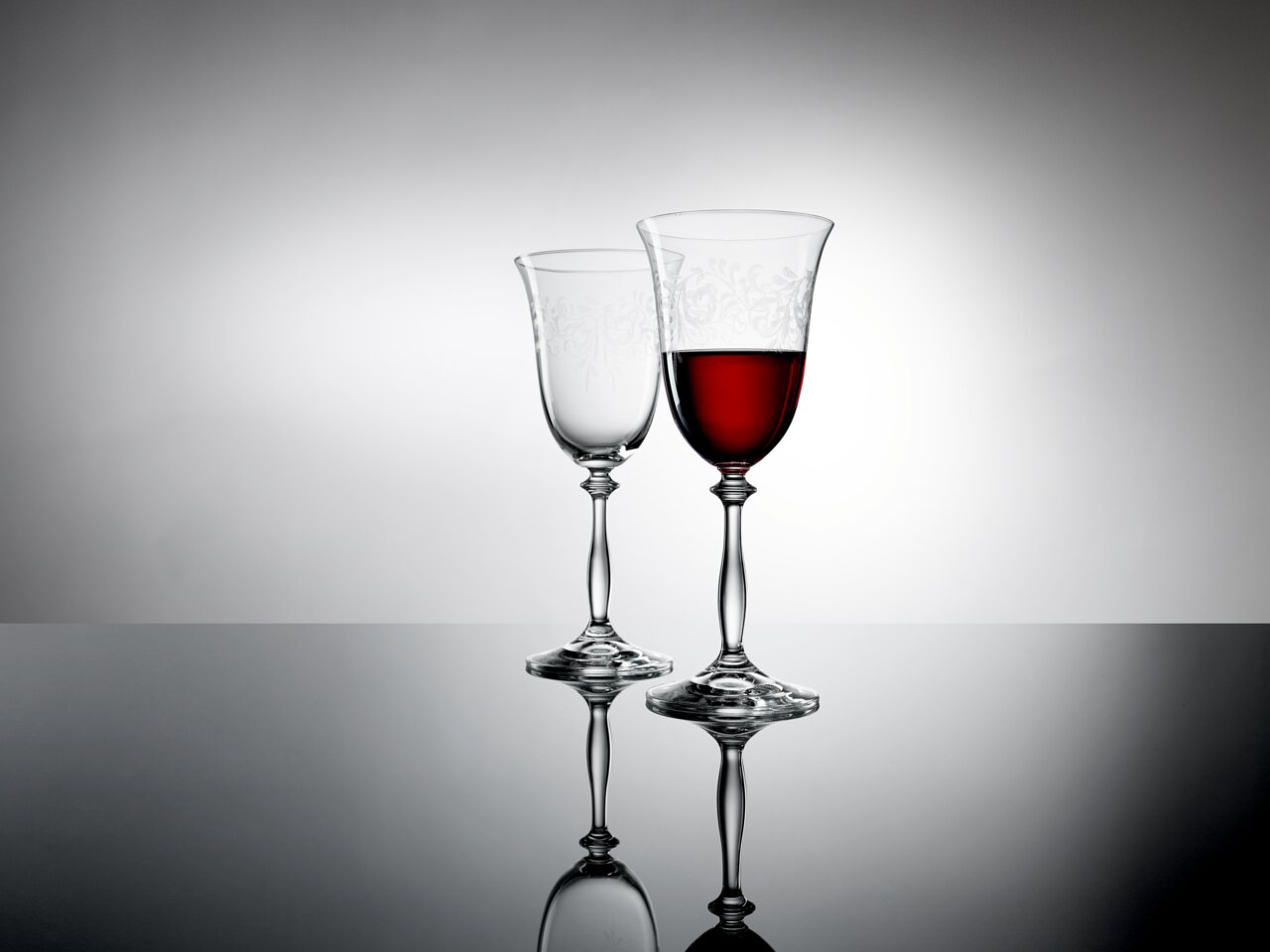 Zwei elegante Weingläser auf einer reflektierenden Oberfläche, eines gefüllt mit Rotwein, seitliche Perspektive.
