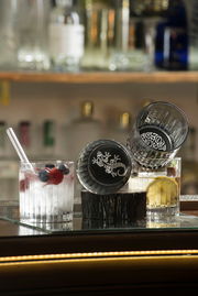 Zwei Whiskygläser mit dekorativem Bodenmuster, eines aufrecht und eines seitlich liegend, gefüllt mit Getränken und Früchten, auf einer Baroberfläche.