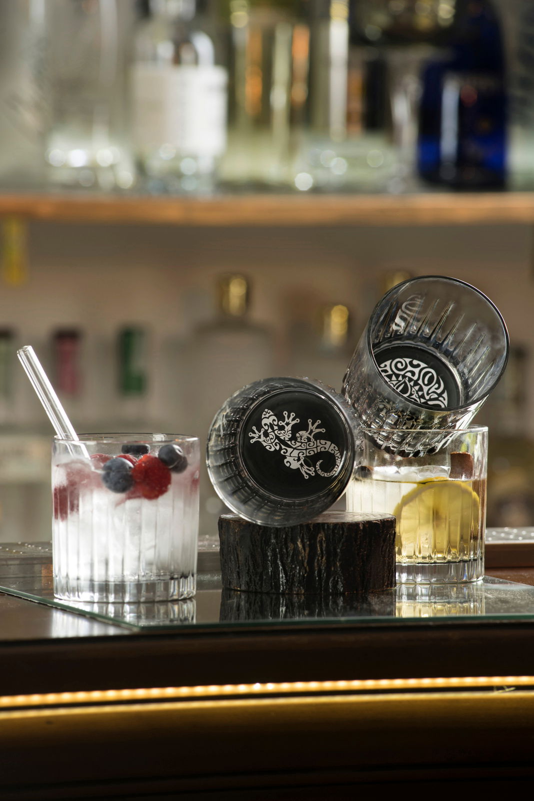 Zwei Whiskygläser mit dekorativem Bodenmuster, eines aufrecht und eines seitlich liegend, gefüllt mit Getränken und Früchten, auf einer Baroberfläche.