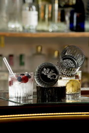 Zwei Whiskygläser mit dekorativem Bodenmuster, eines aufrecht und eines seitlich liegend, gefüllt mit Getränken und Früchten, auf einer Baroberfläche.