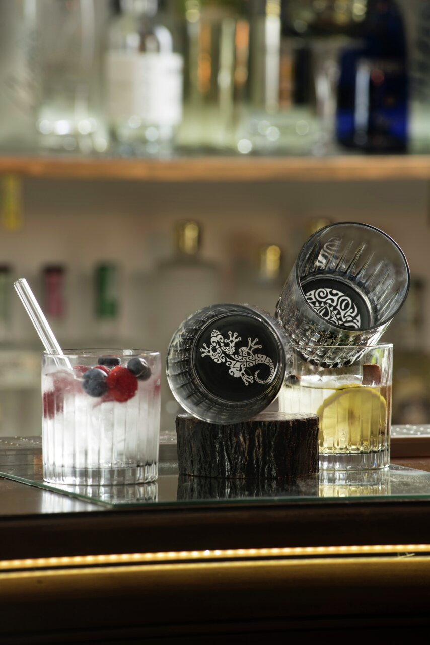 Zwei Whiskygläser mit dekorativem Bodenmuster, eines aufrecht und eines seitlich liegend, gefüllt mit Getränken und Früchten, auf einer Baroberfläche.