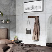 Wandgarderobe aus Holz mit mehreren Haken, an der eine braune Jacke und eine schwarze Tasche hängen, in einem modernen Schlafzimmer mit Betonwänden und minimalistischer Dekoration. Perspektive: seitlich.