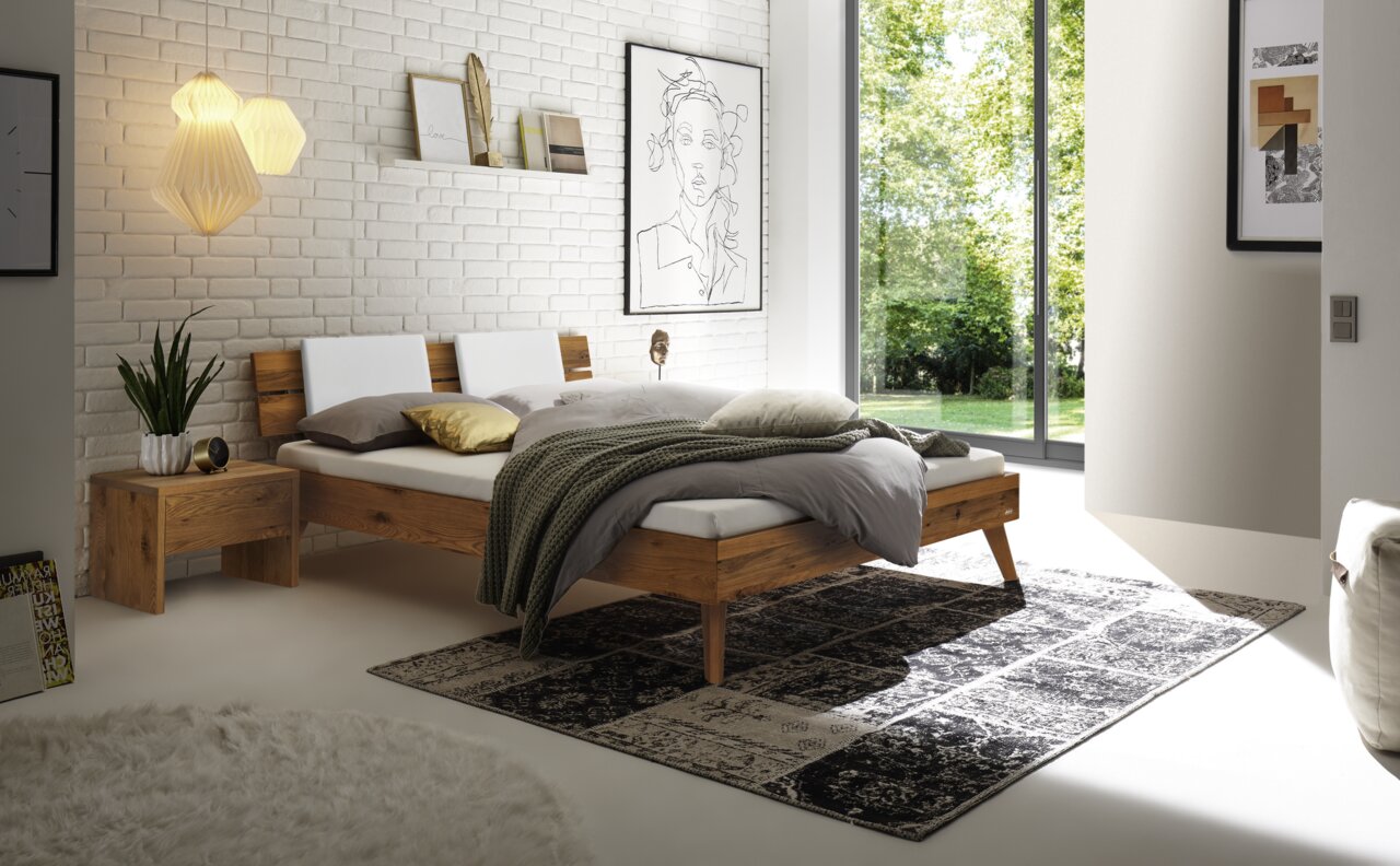 Modernes Schlafzimmer mit einem Kopfteil aus Holz und Metall, seitliche Perspektive. Das Bett ist mit weißen und grauen Kissen sowie einer grünen Decke dekoriert. Ein Nachttisch aus Holz steht daneben, und große Fenster lassen viel Tageslicht herein.