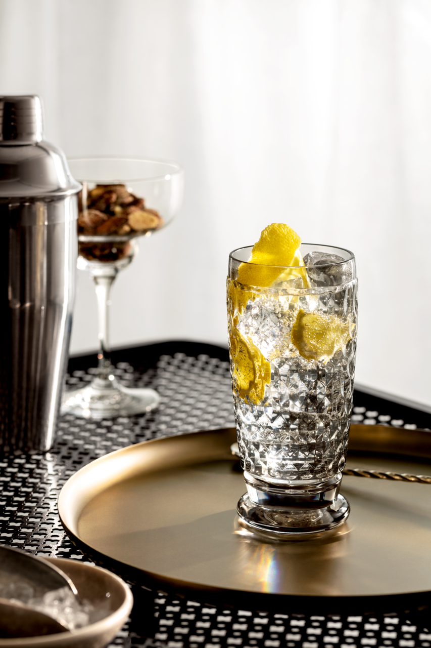 Ein Longdrinkglas mit Eiswürfeln und Zitronenscheiben, auf einem goldenen Tablett stehend, aus seitlicher Perspektive fotografiert.