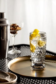 Ein Longdrinkglas mit Eiswürfeln und Zitronenscheiben, auf einem goldenen Tablett stehend, aus seitlicher Perspektive fotografiert.