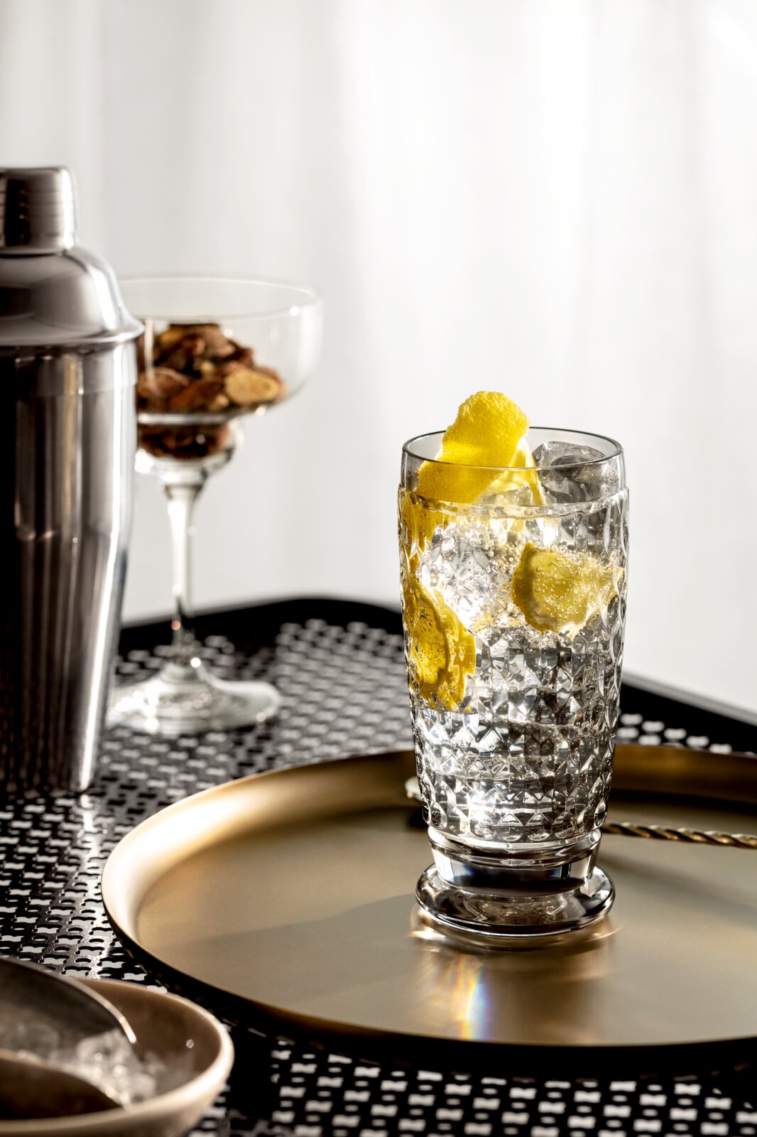 Ein Longdrinkglas mit Eiswürfeln und Zitronenscheiben, auf einem goldenen Tablett stehend, aus seitlicher Perspektive fotografiert.
