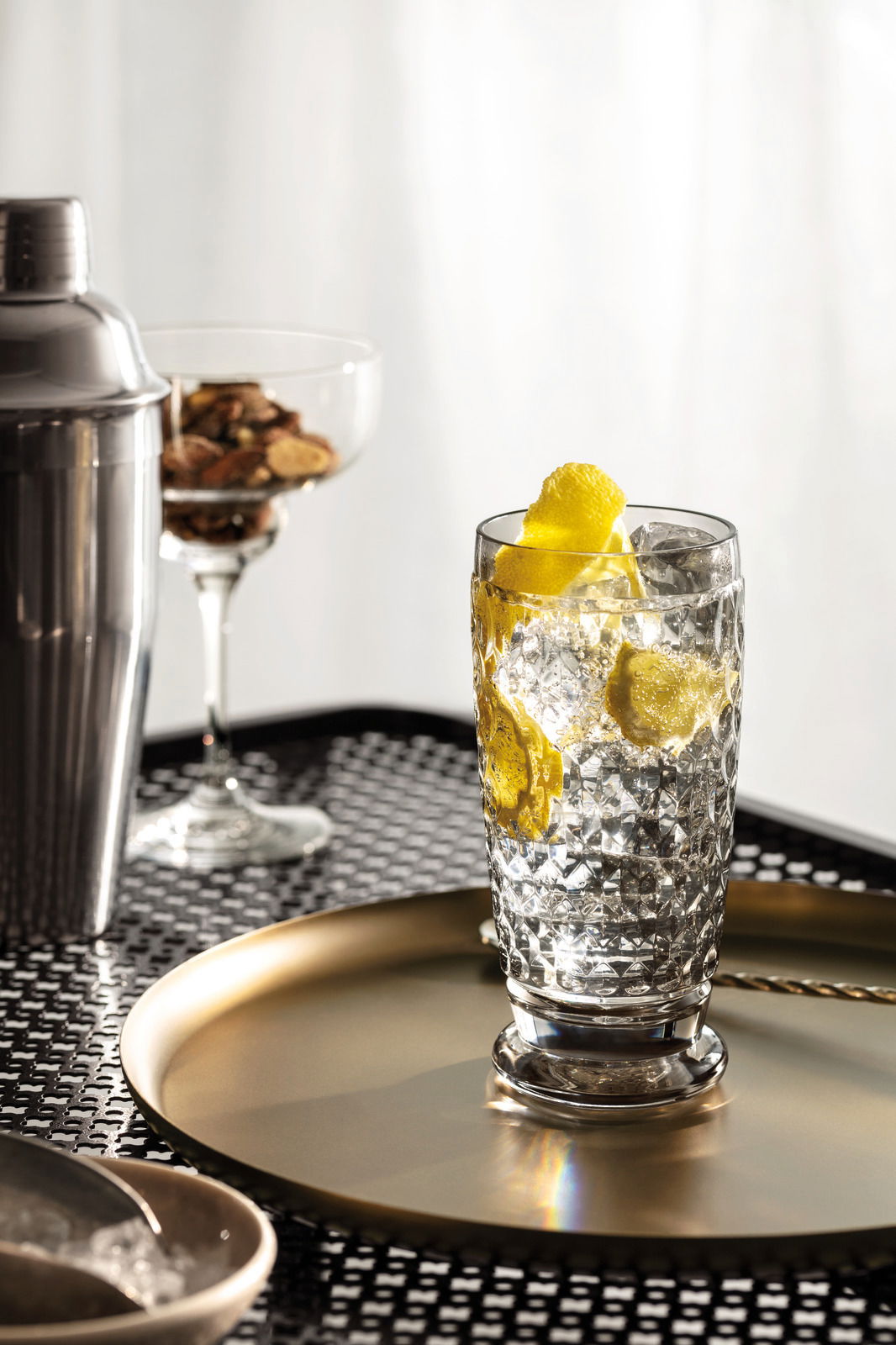 Ein Longdrinkglas mit Eiswürfeln und Zitronenscheiben, auf einem goldenen Tablett stehend, aus seitlicher Perspektive fotografiert.