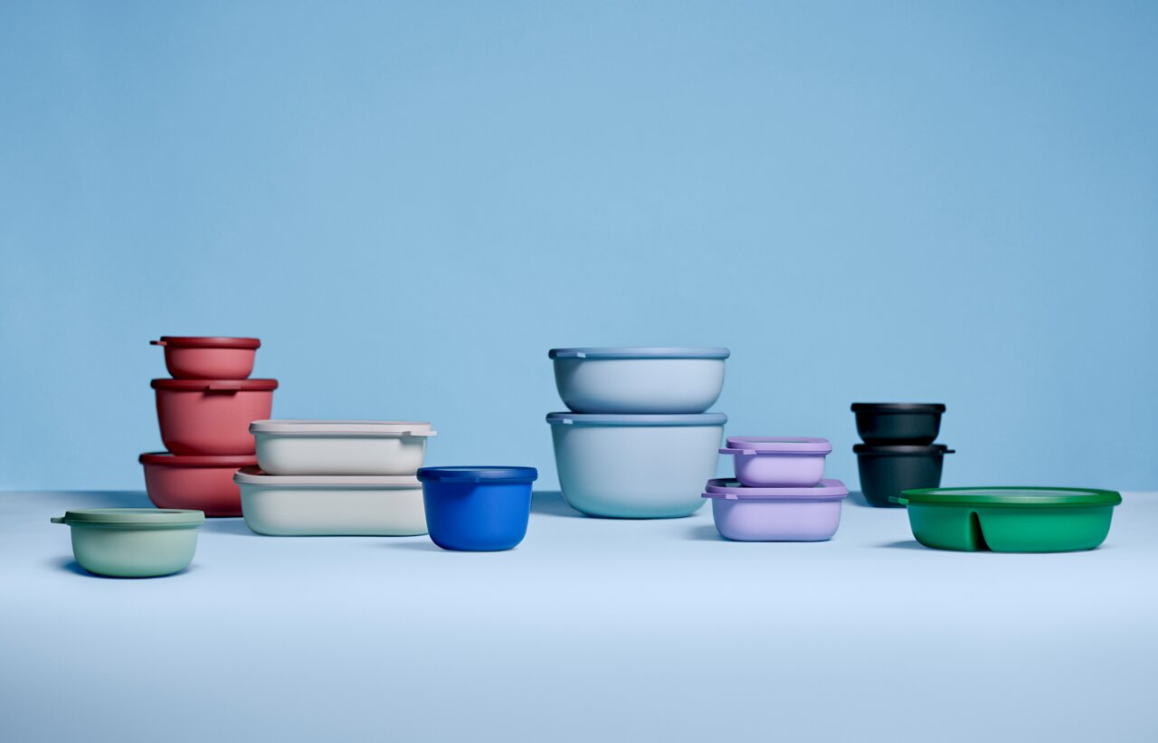 Verschiedene bunte Bento-Schalen in verschiedenen Größen und Farben, darunter rosa, blau, grün und lila, auf einer hellblauen Oberfläche. Die Schalen sind aus Kunststoff und haben runde und rechteckige Formen. Perspektive von vorne.