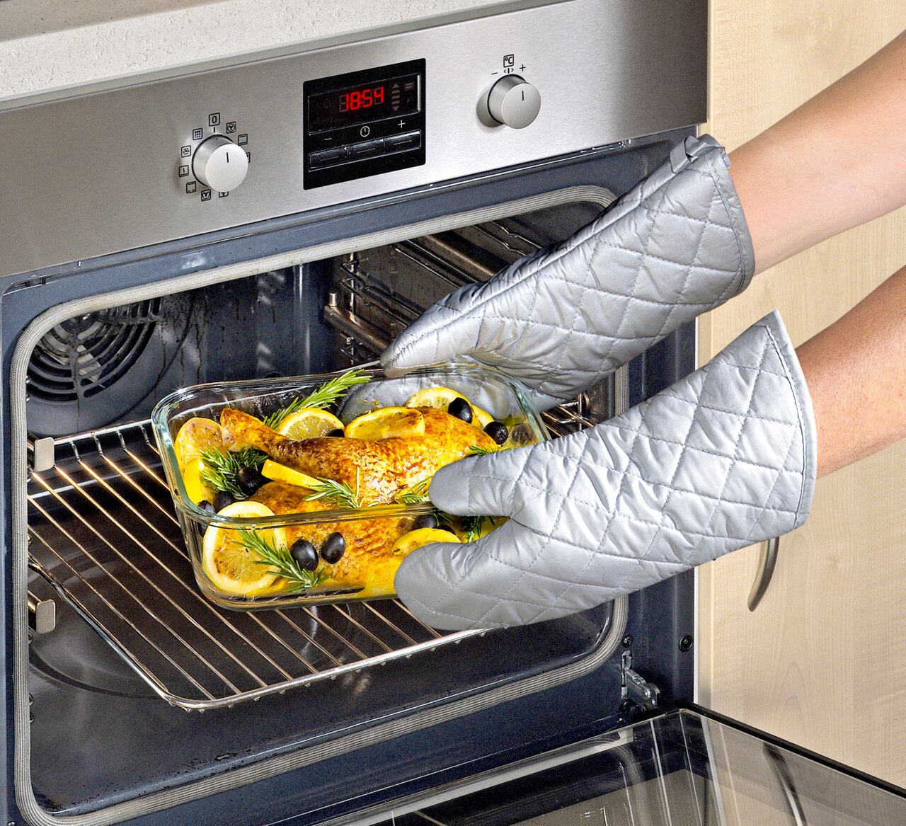 Person mit silbernen Ofenhandschuhen nimmt eine Glasauflaufform mit gebratenem Hähnchen, Zitronenscheiben und Kräutern aus einem modernen Backofen. Perspektive von der Seite.