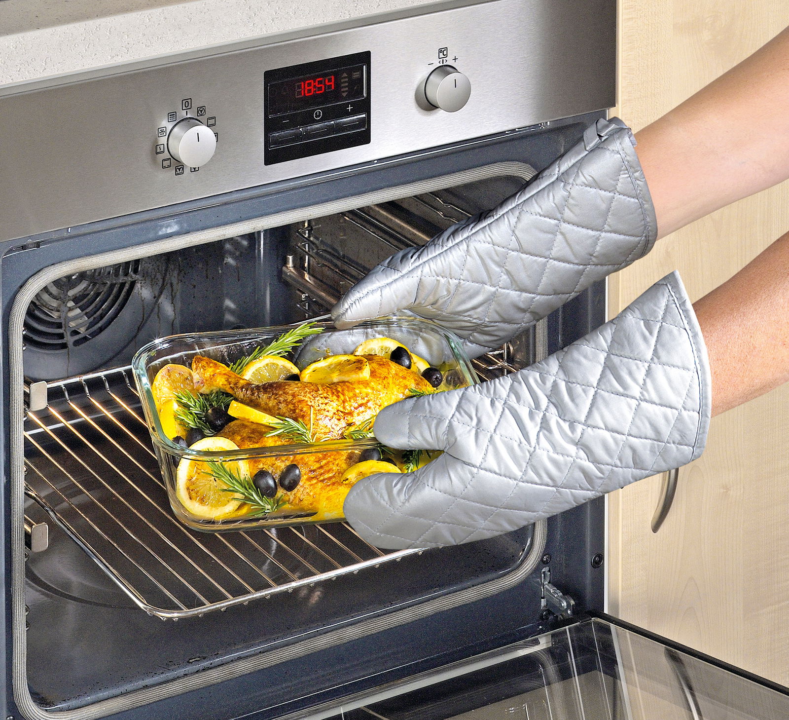 Person mit silbernen Ofenhandschuhen nimmt eine Glasauflaufform mit gebratenem Hähnchen, Zitronenscheiben und Kräutern aus einem modernen Backofen. Perspektive von der Seite.