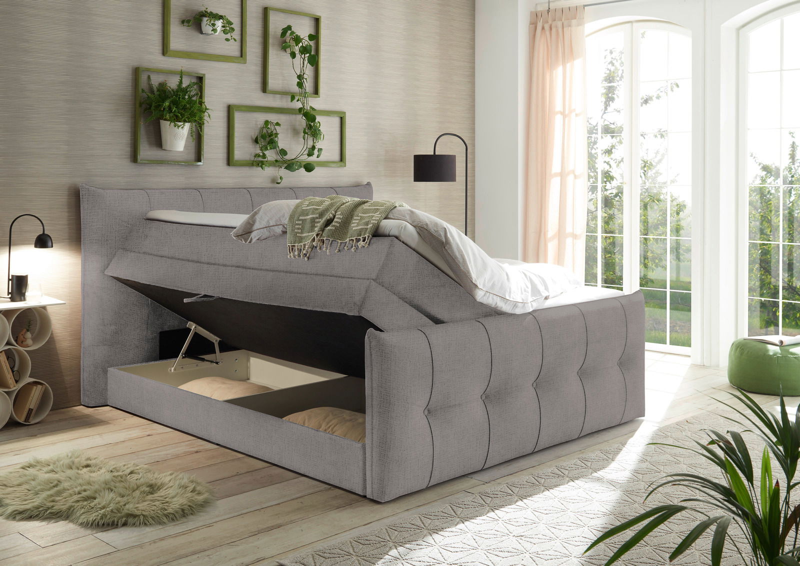 Graues Boxspringbett mit Stauraum, seitliche Perspektive, in einem hellen Raum mit großen Fenstern und Pflanzen.