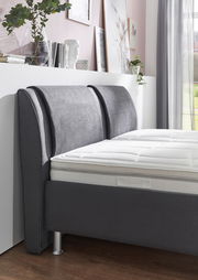 Seitliche Perspektive eines modernen Polsterbetts mit grauem Kopfteil und weißer Matratze, umgeben von stilvoller Dekoration.