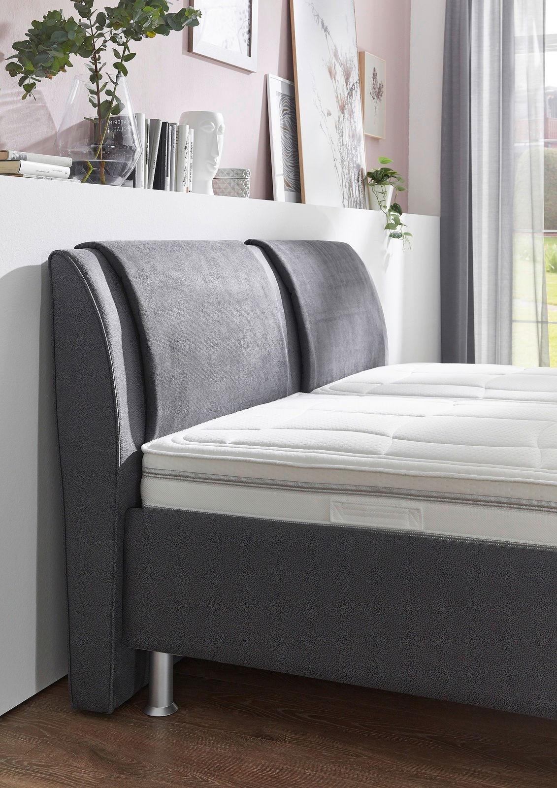 Seitliche Perspektive eines modernen Polsterbetts mit grauem Kopfteil und weißer Matratze, umgeben von stilvoller Dekoration.
