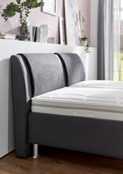 Seitliche Perspektive eines modernen Polsterbetts mit grauem Kopfteil und weißer Matratze, umgeben von stilvoller Dekoration.