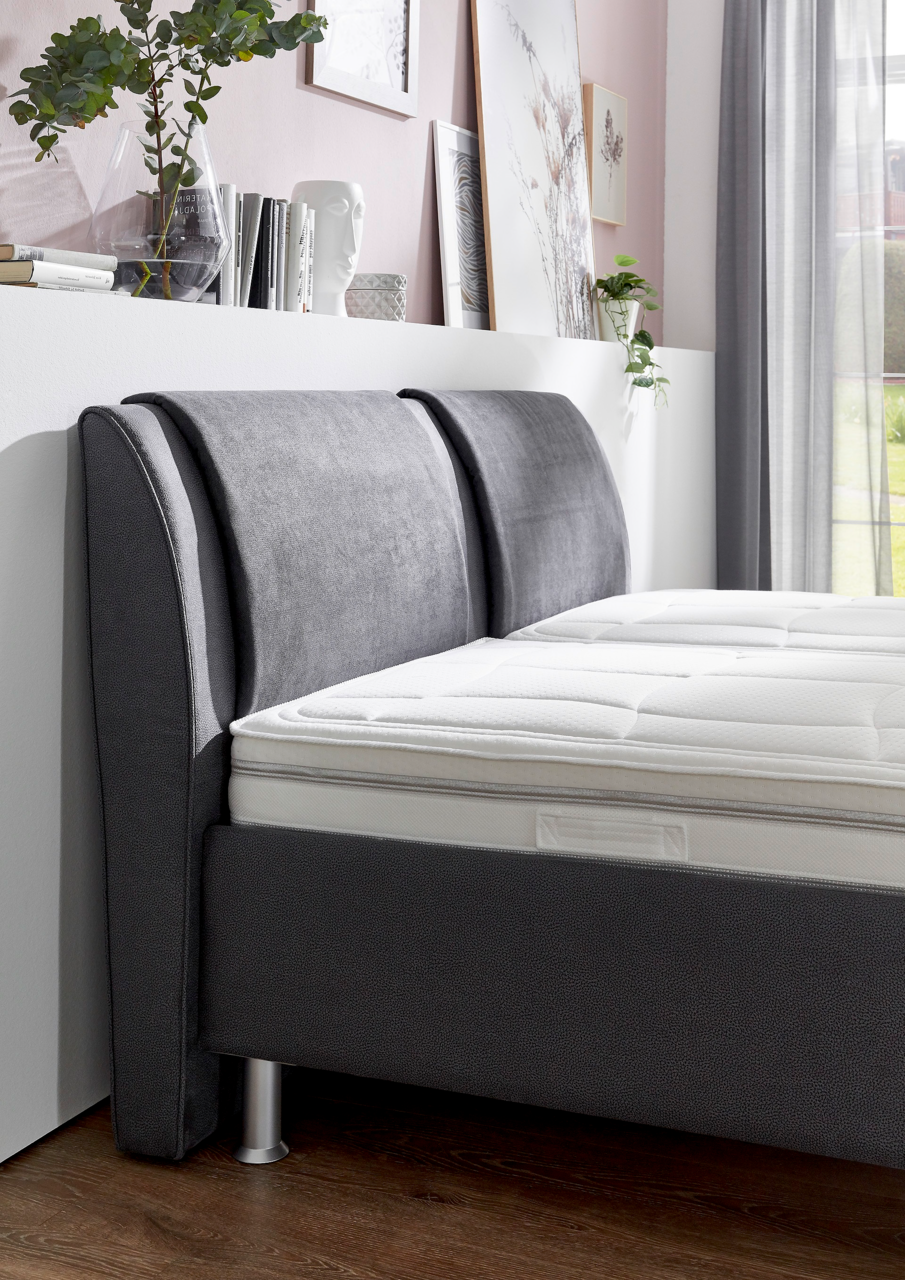 Seitliche Perspektive eines modernen Polsterbetts mit grauem Kopfteil und weißer Matratze, umgeben von stilvoller Dekoration.