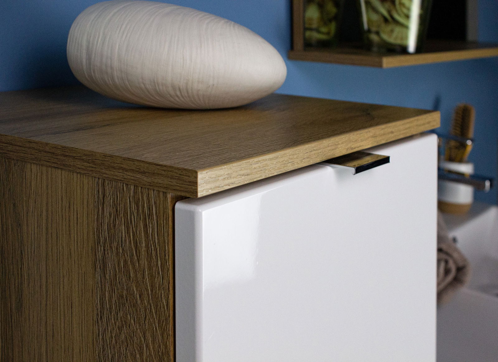 Nahaufnahme eines Schranks mit Holzoberfläche und weißer Tür, darauf liegt eine ovale, weiße Dekoration. Perspektive von schräg oben.