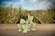 Fünf hellgrüne, gesprenkelte Keramik-Eierbecher auf Holztisch vor Schilflandschaft; Frontansicht auf Augenhöhe, ein Becher liegt seitlich.