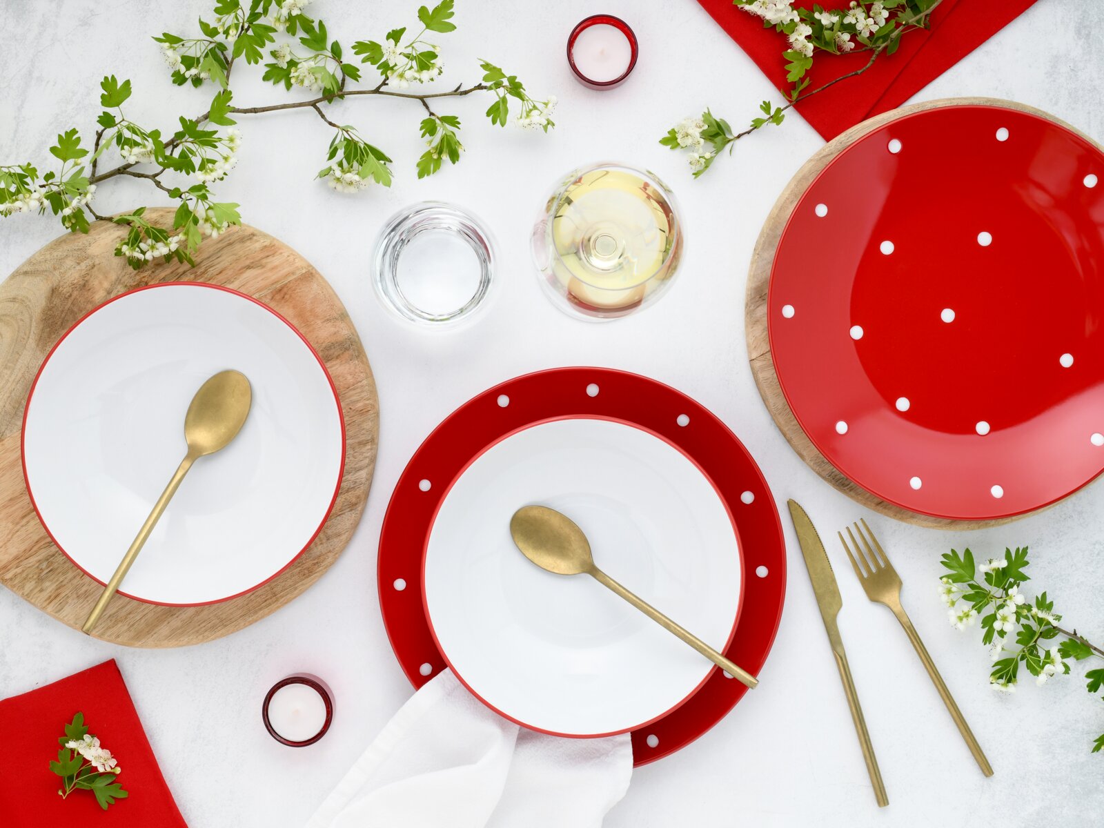 Draufsicht auf ein gedecktes Tisch-Arrangement mit roten, weiß gepunkteten Tellern und weißen Schalen auf Holzuntersetzern, goldfarbenem Besteck, Gläsern, roten Servietten, Teelichtern und blühenden Zweigen auf hellem Hintergrund