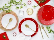 Draufsicht auf ein gedecktes Tisch-Arrangement mit roten, weiß gepunkteten Tellern und weißen Schalen auf Holzuntersetzern, goldfarbenem Besteck, Gläsern, roten Servietten, Teelichtern und blühenden Zweigen auf hellem Hintergrund
