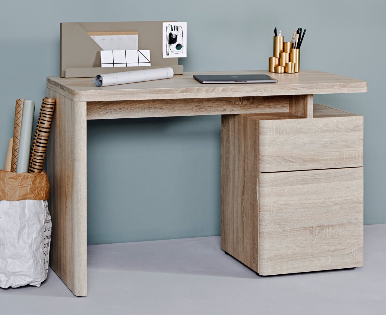 Moderner Schreibtisch aus hellem Holz mit Schubladen, seitlicher Perspektive. Dekoriert mit Laptop, Stiftehalter und Papierrollen.