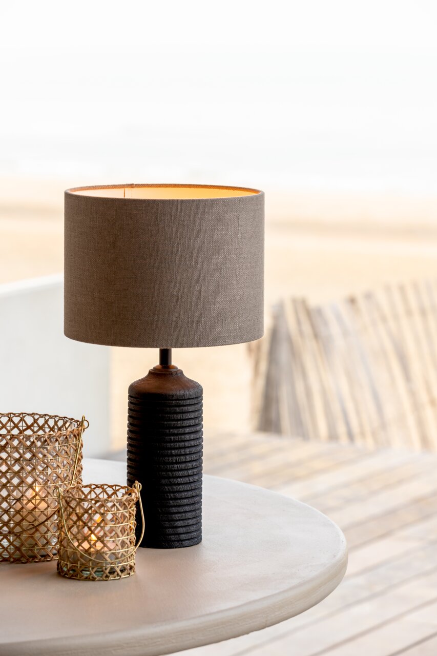 Zylindrischer Lampenschirm in neutralem Beige auf einem Tisch, mit einem dekorativen, geriffelten Lampenfuß, seitliche Perspektive.