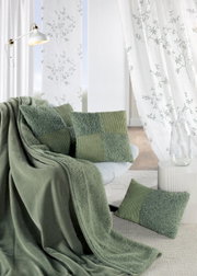 Gemütliche Wohnzimmerszene mit grünen Kissen und Decke auf einem Sofa, umgeben von weißen Vorhängen mit Pflanzenmuster. Perspektive von vorne.