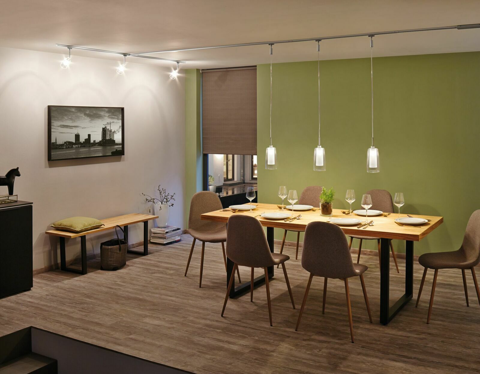 Esszimmer mit modernem Schienenpendel Arido II, das über einem Esstisch mit sechs Stühlen hängt. Die Perspektive zeigt den Raum von der Seite, mit einem grünen Akzent an der Wand.