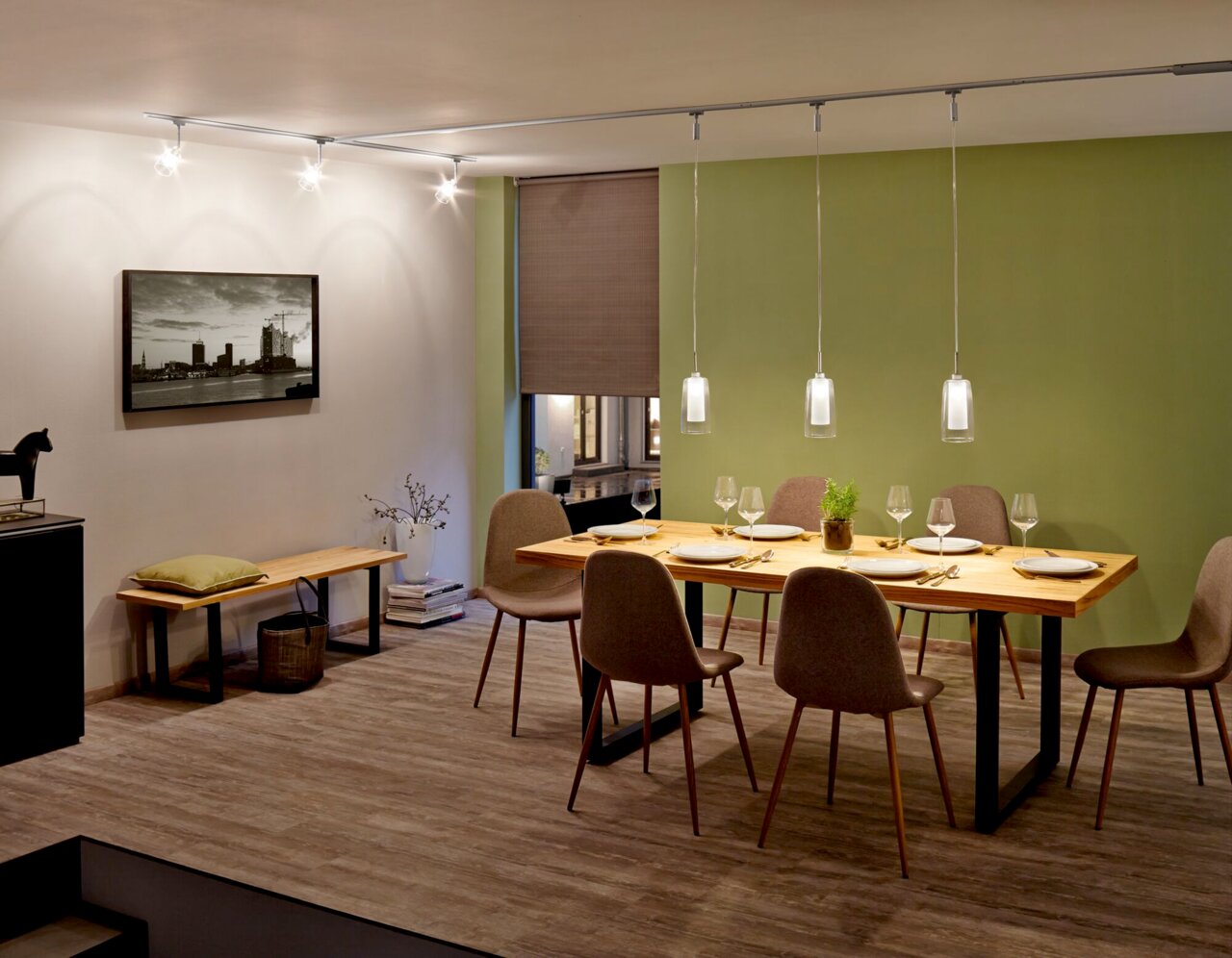 Esszimmer mit modernem Schienenpendel Arido II, das über einem Esstisch mit sechs Stühlen hängt. Die Perspektive zeigt den Raum von der Seite, mit einem grünen Akzent an der Wand.