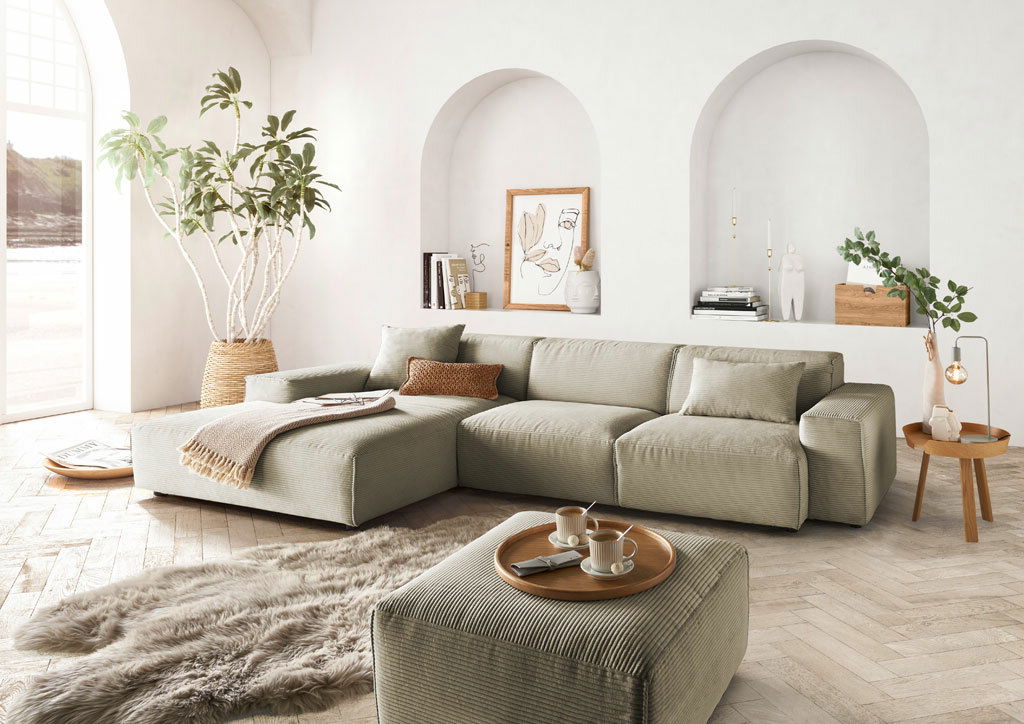 Lounge Handmade Ecksofa LH-EDINA Moderne Rundecke in einem hellen Wohnzimmer mit großen Fenstern, Pflanzen und stilvollen Dekorationen, aus der Perspektive von vorne links.