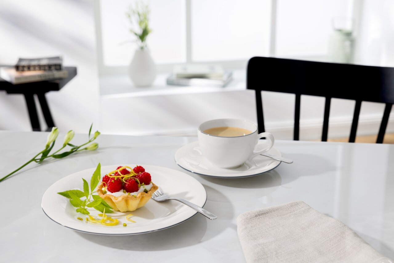 Kaffeetasse auf einem Tisch mit einem Teller mit Himbeertörtchen, von schräg oben fotografiert.